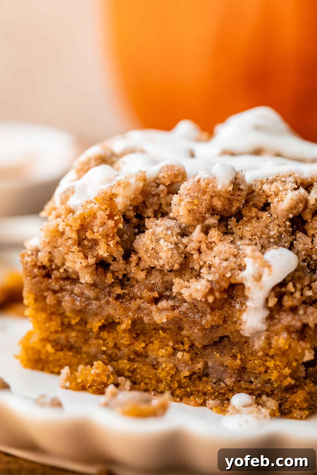 Spiced Pumpkin Crumble Coffee Cake 19 A beautiful slice of pumpkin streusel coffee cake on a white scalloped plate, with decorative pumpkins softly blurred in the background, creating a festive autumn scene.
