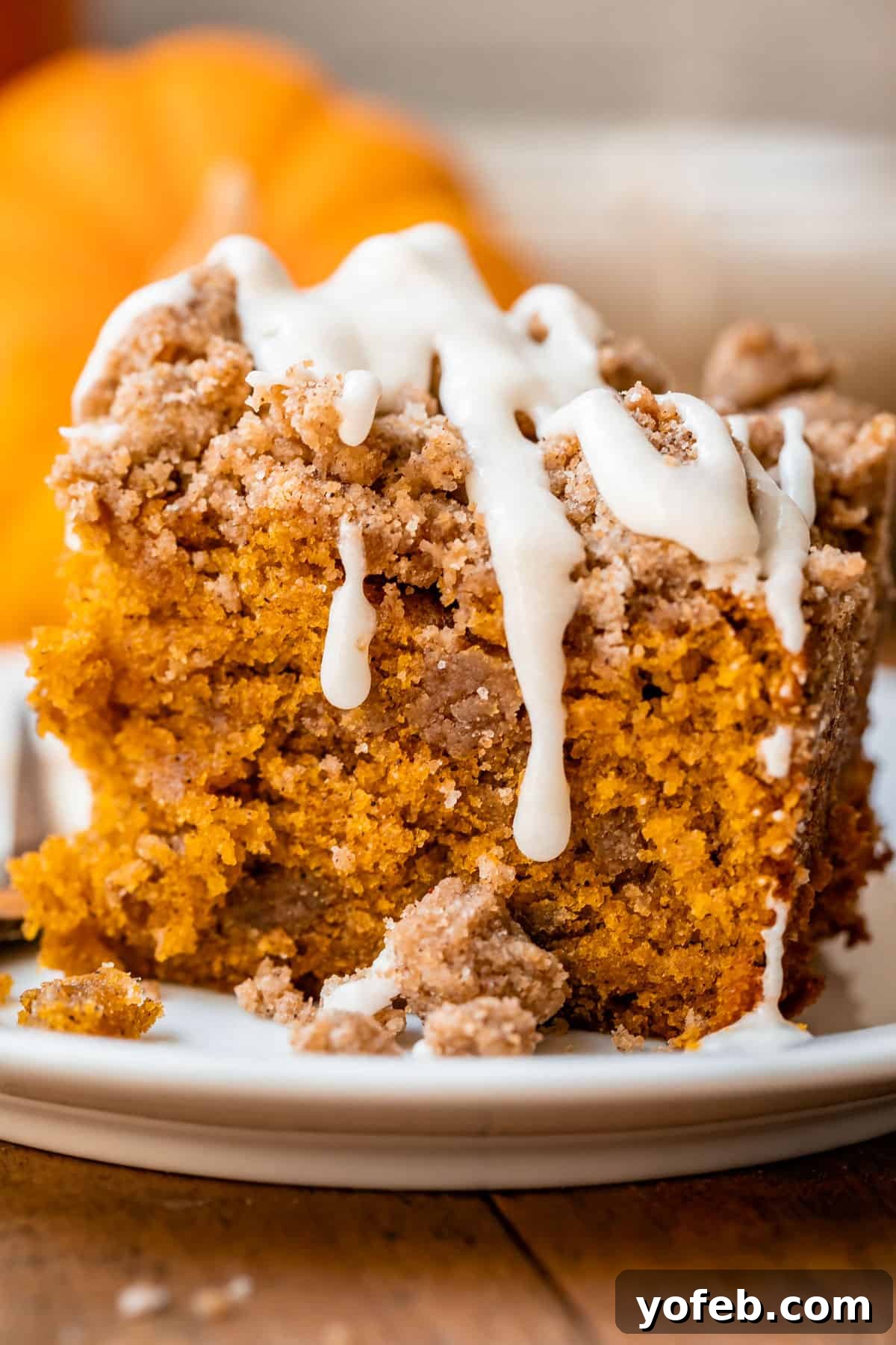 Spiced Pumpkin Crumble Coffee Cake 21 A slice of pumpkin spice coffee cake on a white plate, with decorative pumpkins softly blurred in the background, creating a cozy and inviting autumnal setting.