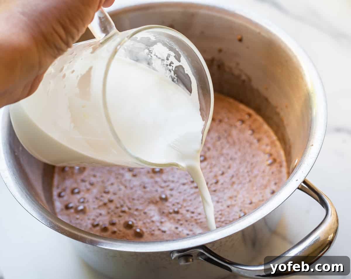 Decadent Chocolate Cream Pie Perfection 17 A hand pouring hot milk and cream mixture into a pot containing beaten eggs and milk, using a hand mixer on low speed to combine.