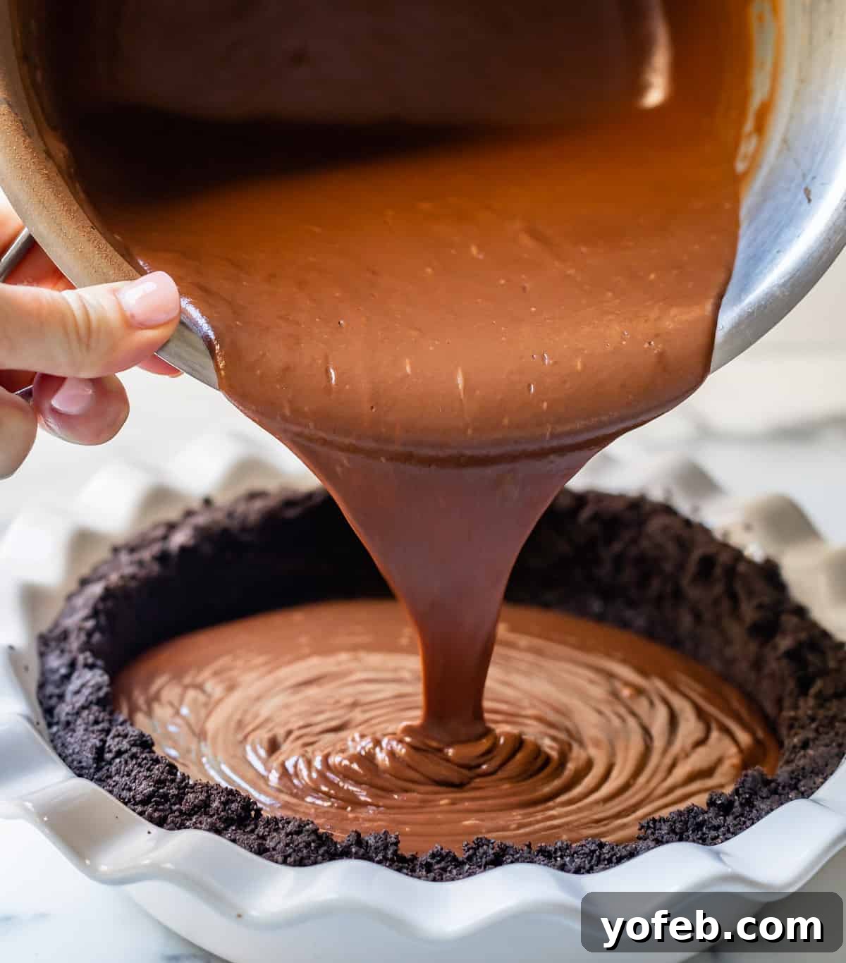 Decadent Chocolate Cream Pie Perfection 22 Rich, dark chocolate pudding being carefully poured into a baked Oreo pie crust in a white ceramic dish.