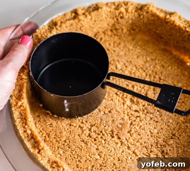 Decadent Chocolate Cream Pie Perfection 10 A black measuring cup being used to press graham cracker crumbs firmly into a pie pan, creating an even crust.