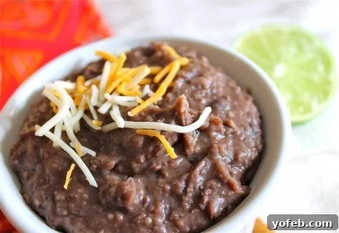 A bowl of slow cooker refried beans topped with shredded cheese, served with a slice of lime and a red napkin in the background.