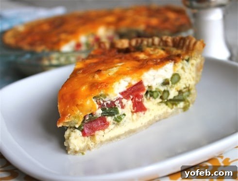 Sun-Kissed Tomato and Asparagus Quiche with Creamy Chèvre 5 Slice of quiche with tomato, asparagus, and goat cheese on a white plate with whole quiche and salt shaker in the background.