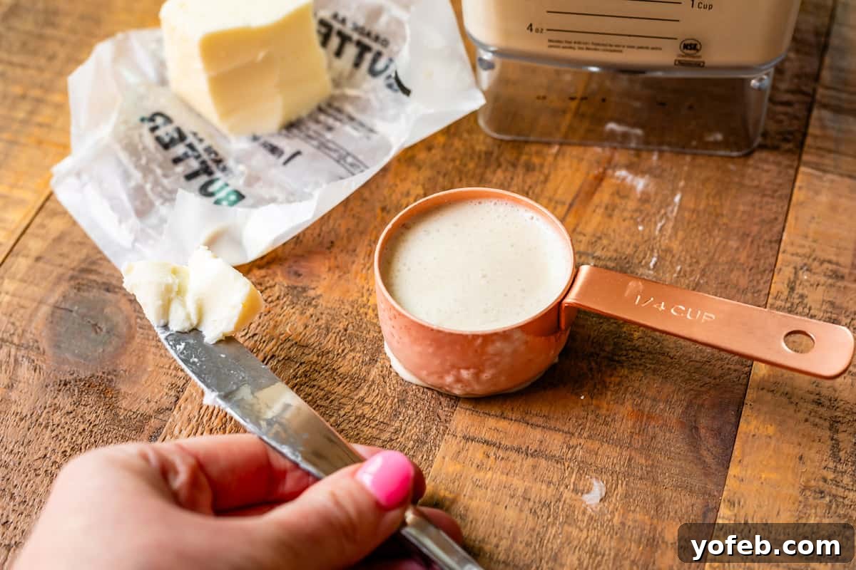 Absolutely The Best Crepes 13 A hand holding a knife with a pat of butter, positioned over a countertop next to a measuring cup filled with crepe batter, setting the scene for cooking.