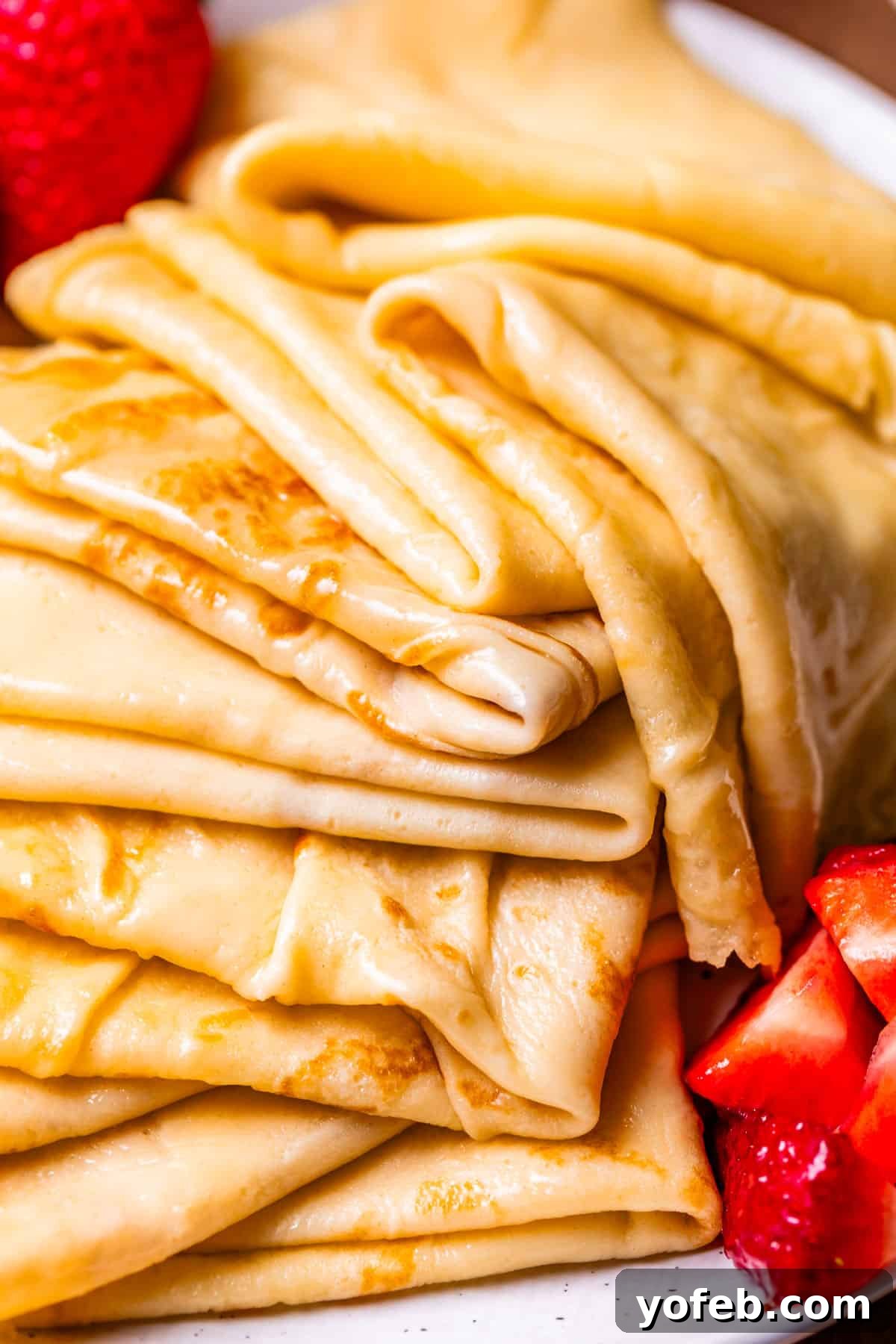 Absolutely The Best Crepes 3 A generous stack of golden-brown crepes, folded into quarters, elegantly arranged beside a pile of fresh, ripe strawberries. A perfect combination for a delightful breakfast or dessert.