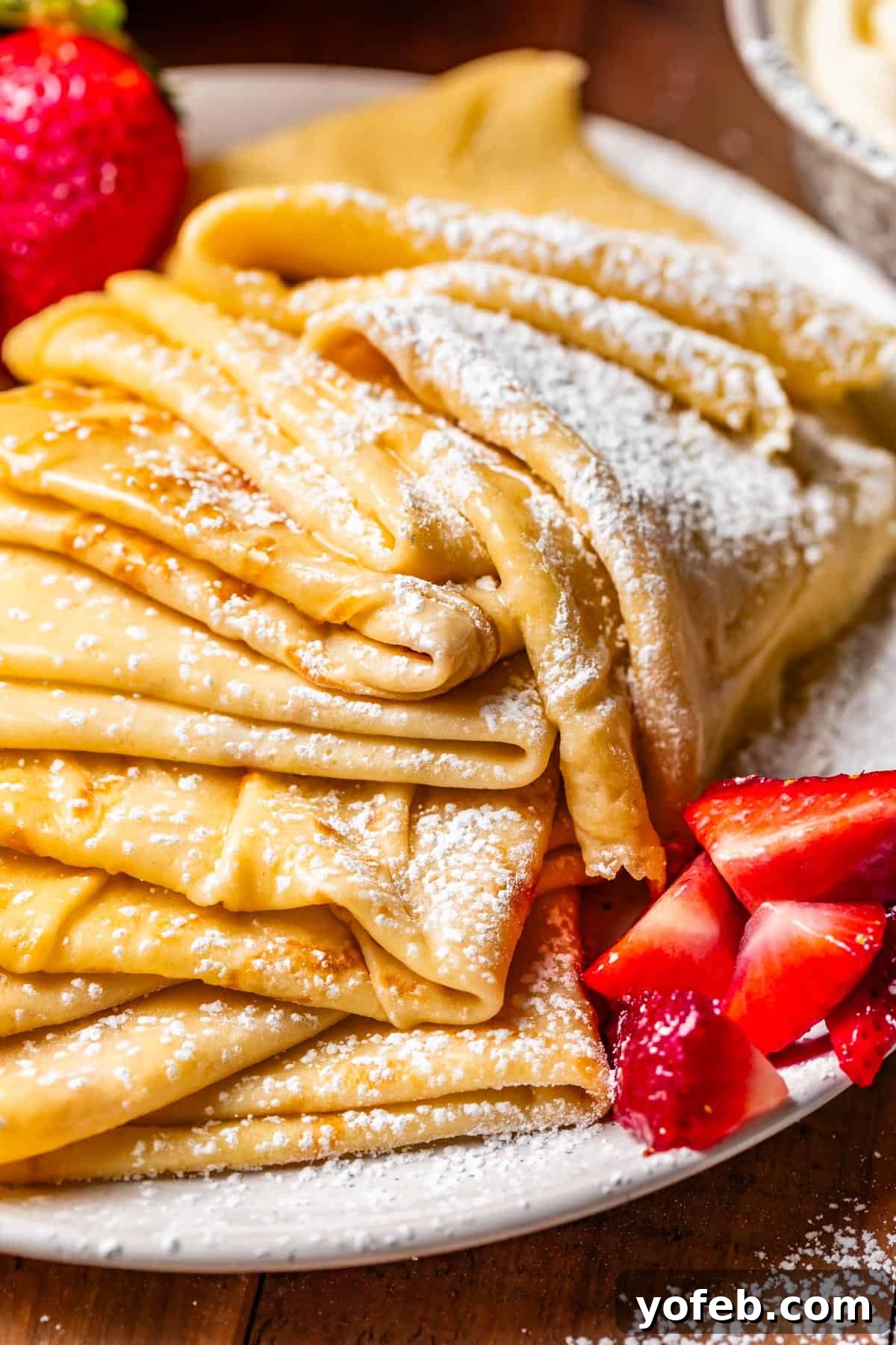 Absolutely The Best Crepes 25 A large, inviting stack of freshly folded crepes, generously dusted with powdered sugar, with a vibrant side of whole strawberries. A visually appealing spread for a delicious breakfast.