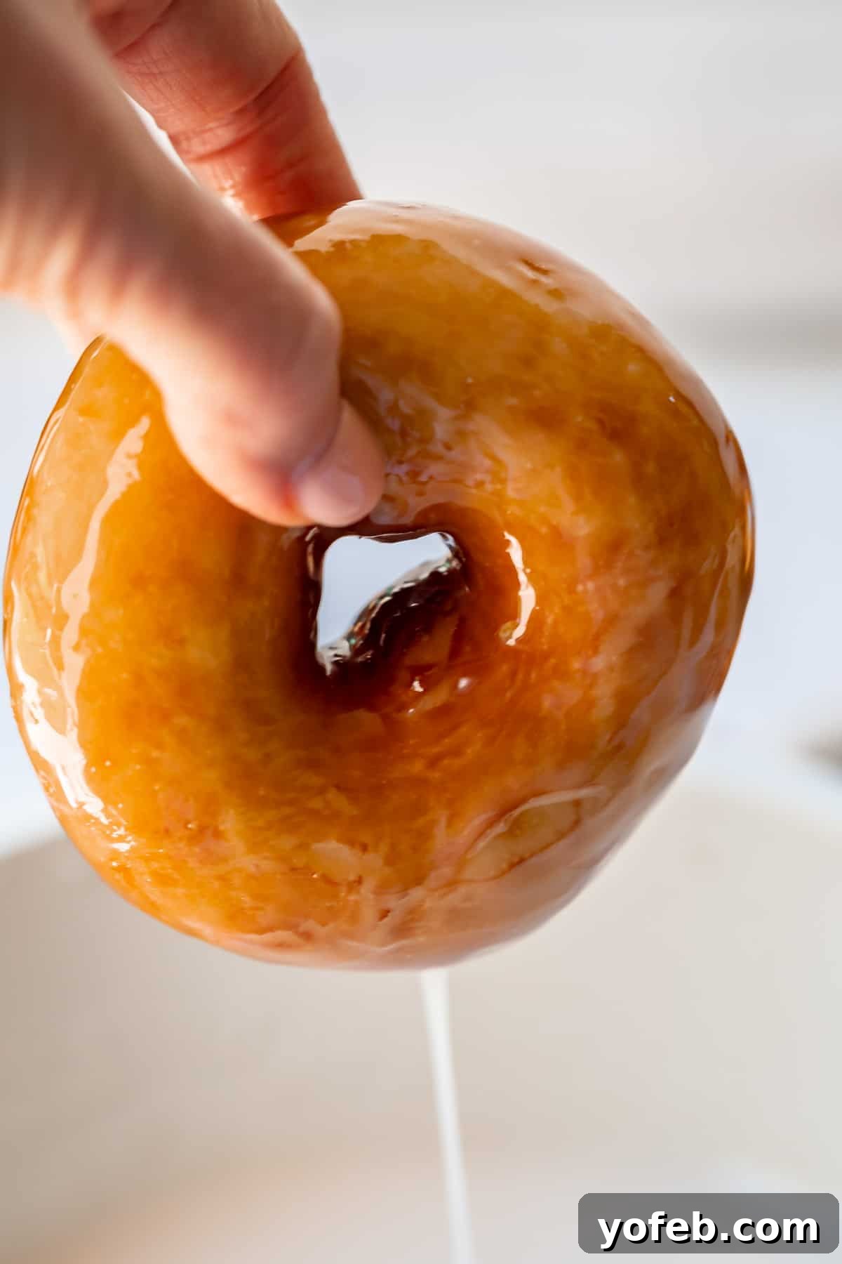 Doughnut Delights from Scratch 35 Fingers carefully pulling a fully coated, glazed donut out of the glaze bowl, allowing excess glaze to drip off before setting.