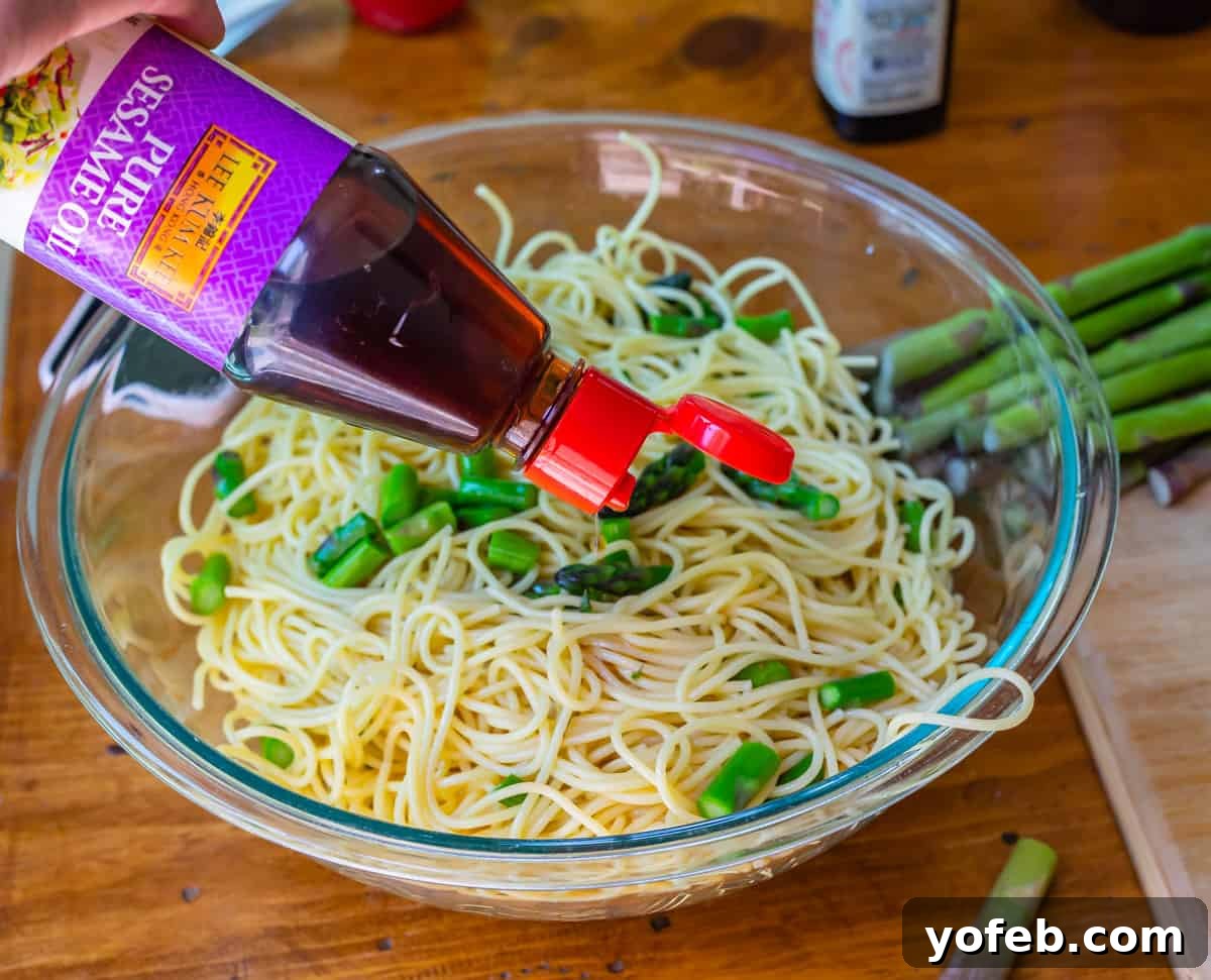 Vibrant Spring Sesame Noodles 11 Sesame oil being drizzled from a bottle over cooked and cooled noodles and vegetables in a large bowl.