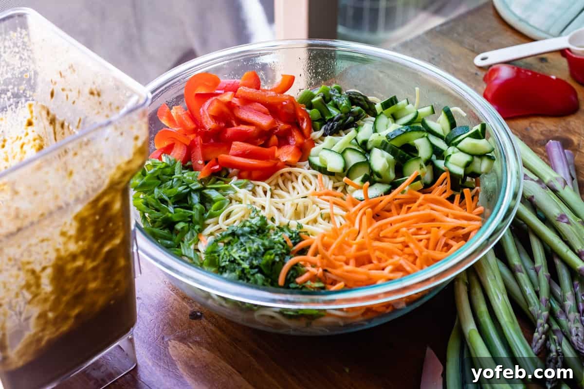 Vibrant Spring Sesame Noodles 15 Chopped bell peppers, cucumber, carrots, cilantro, green onion, and asparagus mixed with cooked noodles in a large glass bowl, before adding sauce.