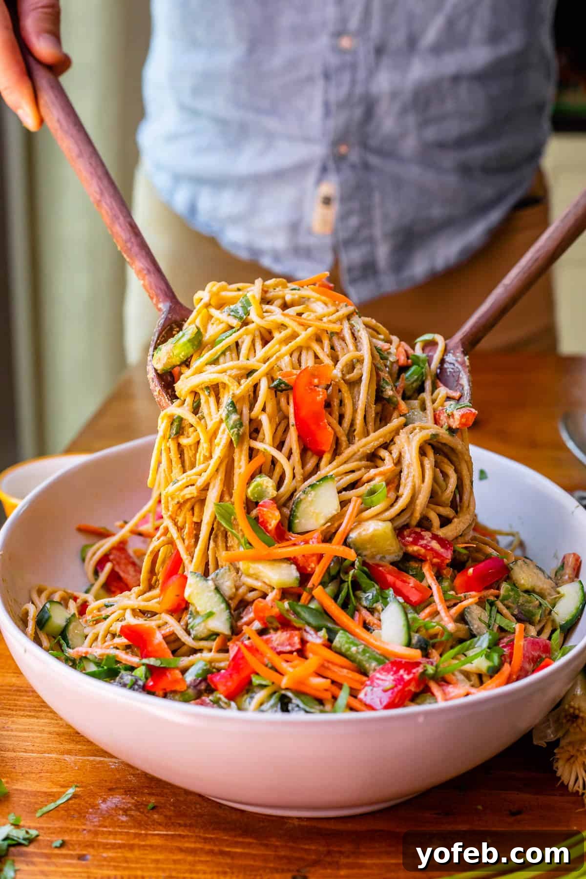 Vibrant Spring Sesame Noodles 18 A ceramic bowl filled with cold sesame noodles and mixed vegetables, being served with two wooden salad spoons.