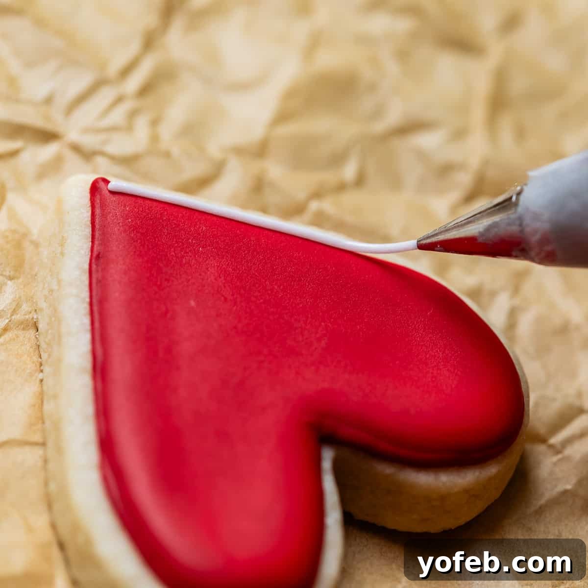 Beginner's Guide to Perfect Royal Icing 27 close up shot of a heart shaped cookie with red frosting on it, being lined with white piping.