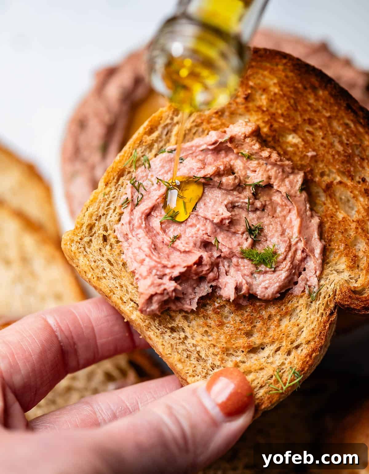 A hand gently drizzles olive oil over a generous spread of liver pâté on toasted rye bread, enhancing its inviting aroma and texture.