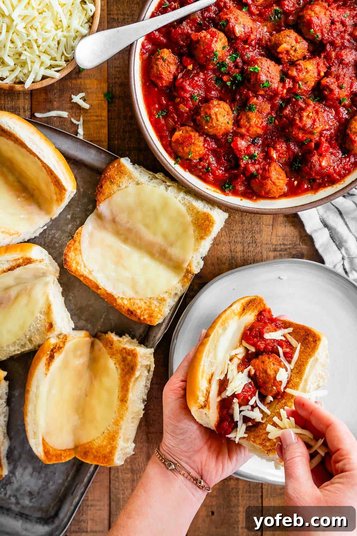 Laura's Ultimate Meatball Sub 11 Hands carefully sprinkling an additional layer of shredded cheese over a generous filling of meatballs and sauce, nestled in a toasted hoagie roll that already has a melted cheese barrier.