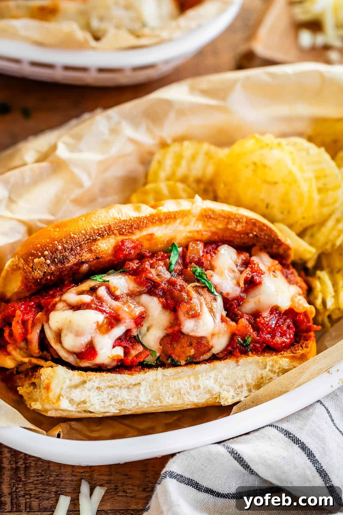 Laura's Ultimate Meatball Sub 13 A delectable meatball sub, garnished with a sprig of fresh basil and served alongside a handful of crispy potato chips, presented in an oval plastic bowl with parchment paper for a classic diner feel.