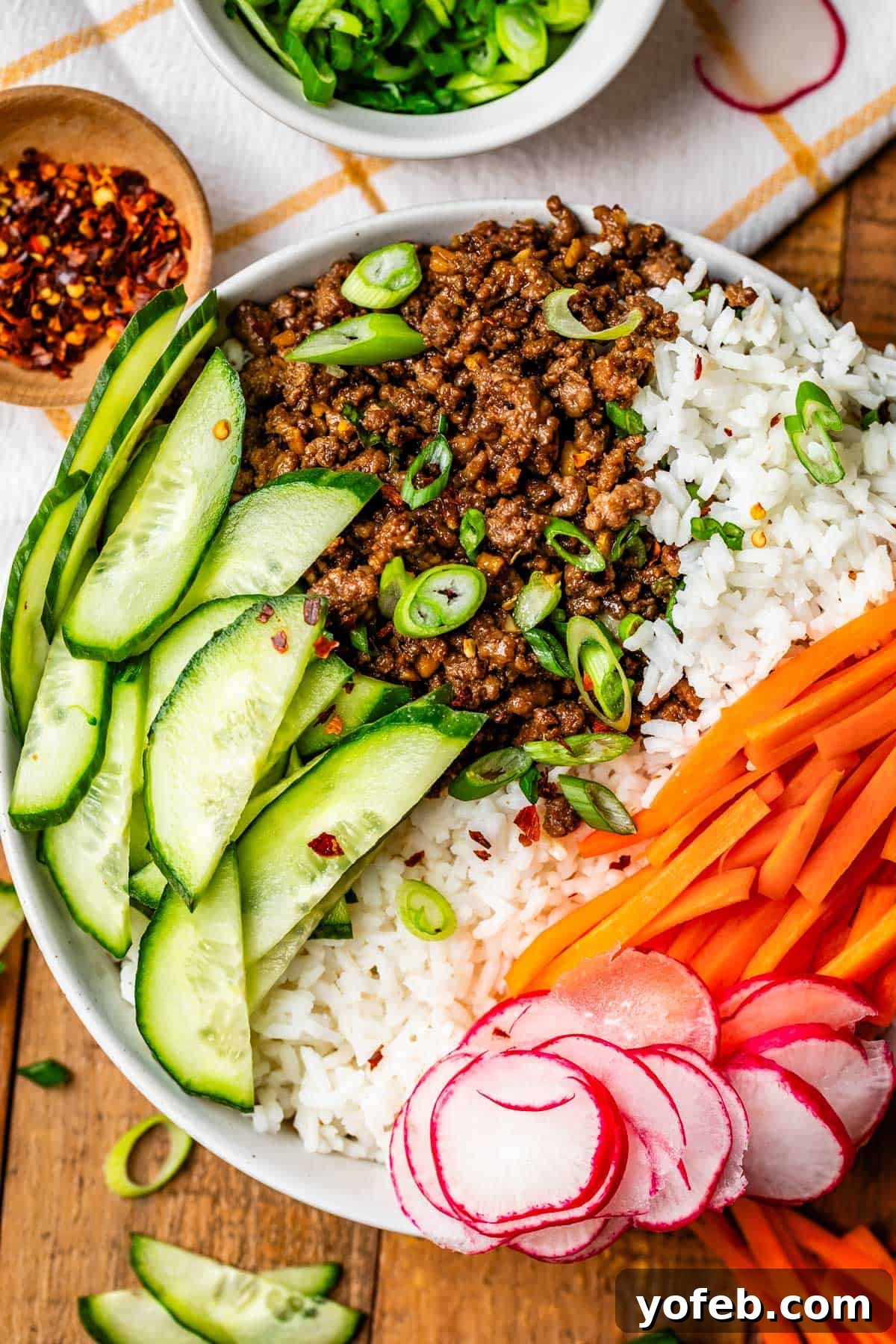 Easy 30-Minute Korean Beef 13 A Korean beef bowl with rice and fresh vegetables sits on a wooden table, next to a small bowl of red pepper flakes for extra spice.