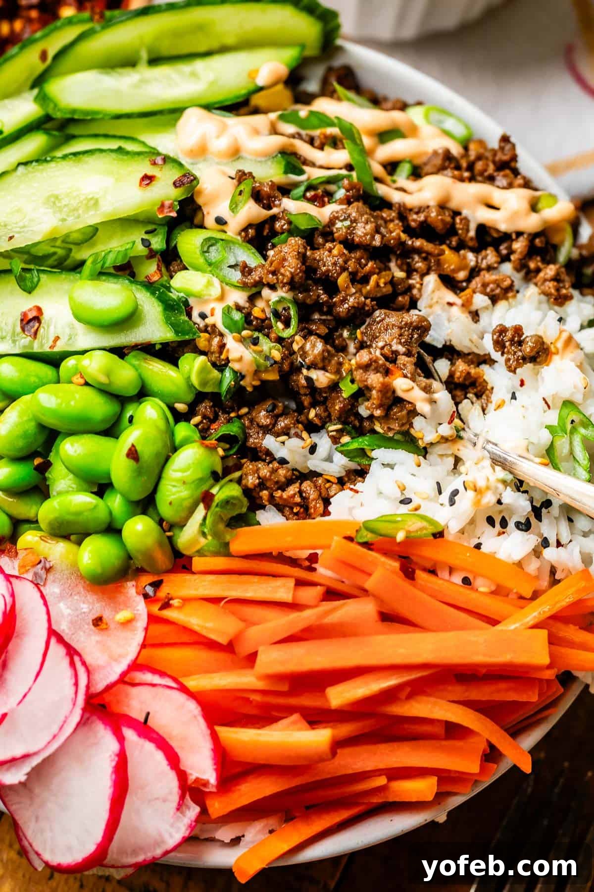 Easy 30-Minute Korean Beef 16 A spoon digging into a colorful Korean beef bowl, revealing layers of rice, savory beef, green edamame, crisp carrot sticks, cool cucumber, and more.