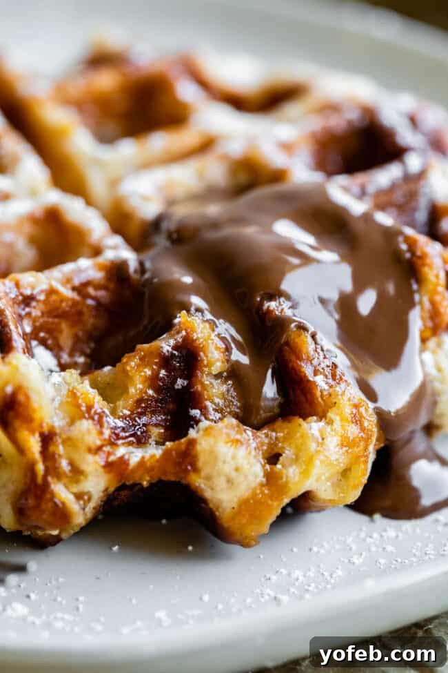 A close-up of a golden brown Liege waffle topped with melted chocolate sauce on a white plate, lightly sprinkled with powdered sugar.
