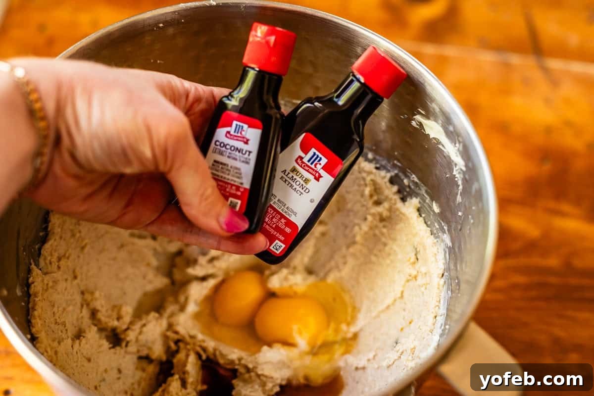 No Roll Chewy Sugar Cookies 30 Minute Recipe 11 A hand holding bottles of coconut and almond extract over a mixing bowl containing cookie dough.