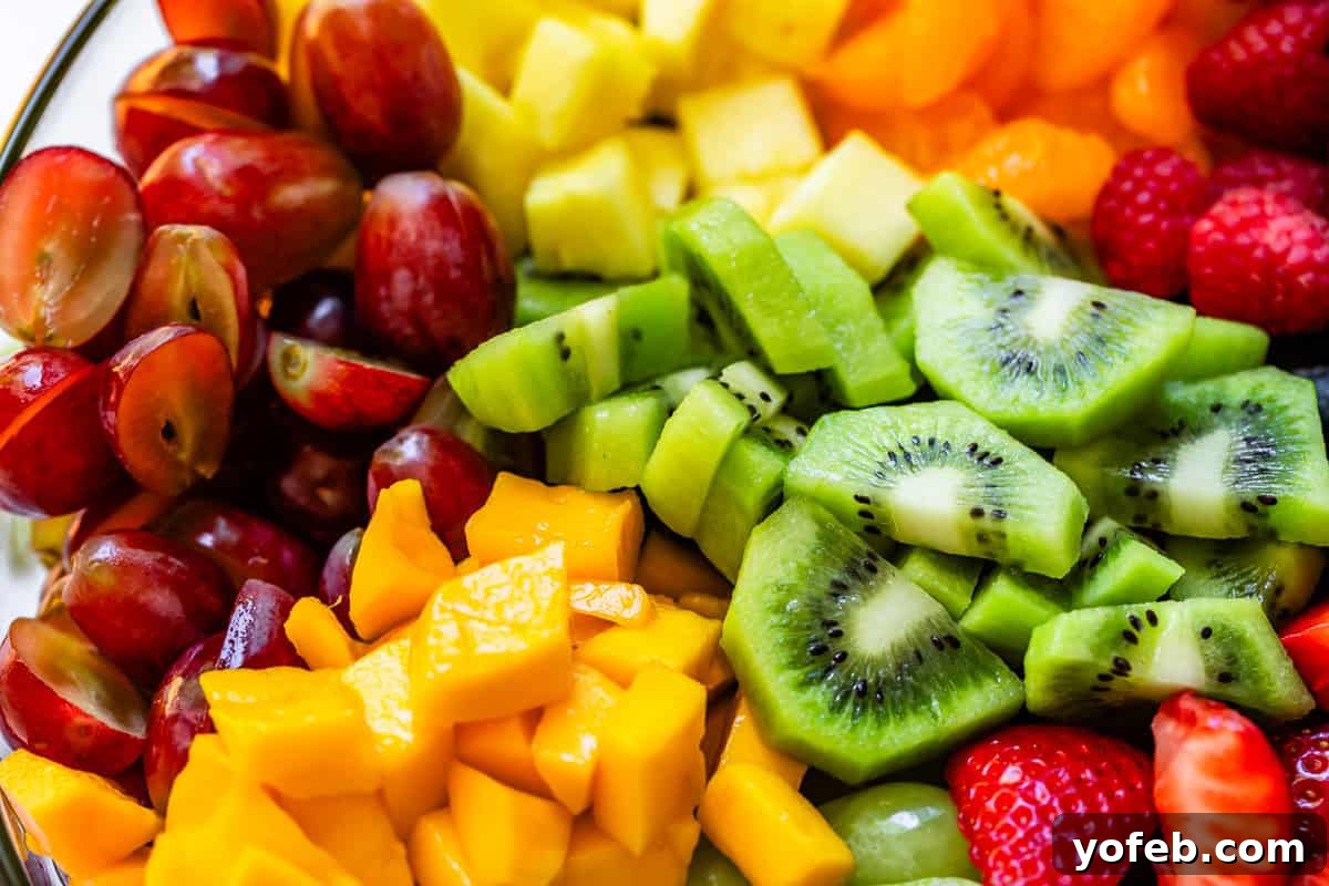 A vibrant close-up of a meticulously cut fruit salad, featuring perfectly diced kiwi, mango, and pineapple, alongside halved strawberries, grapes, and plump raspberries, all ready for the final toss.
