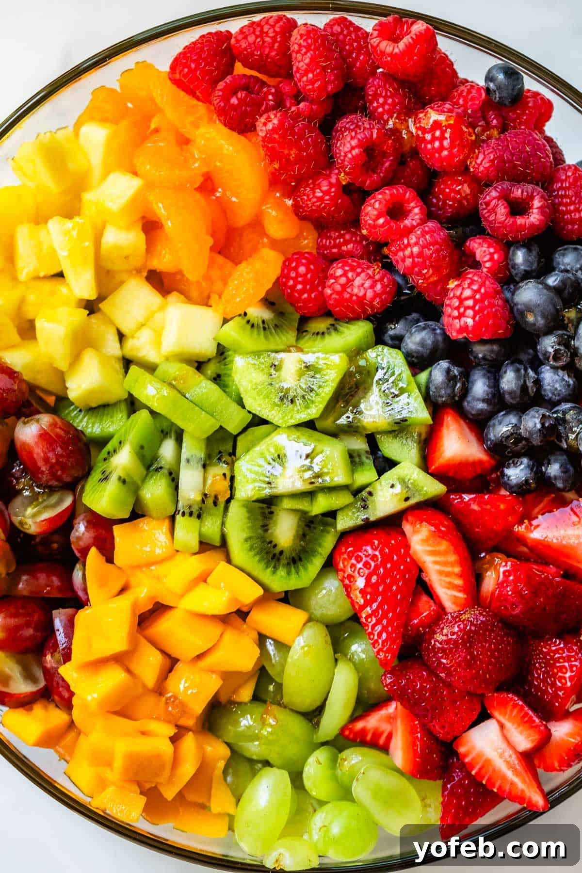 A pristine glass bowl filled with an unmixed assortment of fresh berries, vibrant kiwi, tropical pineapple, juicy mango, and plump grapes, all ready for the final touch of glaze and gentle stir.