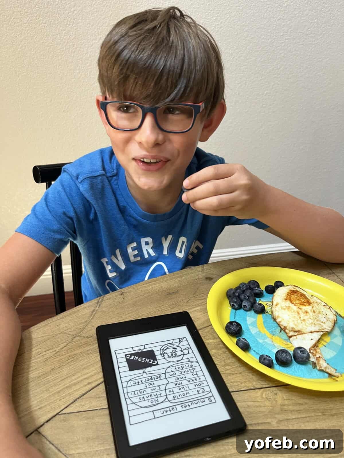 A young boy, Truman, contentedly eating blueberries and an egg while engrossed in reading a Kindle, illustrating his preference for healthy, fresh foods.