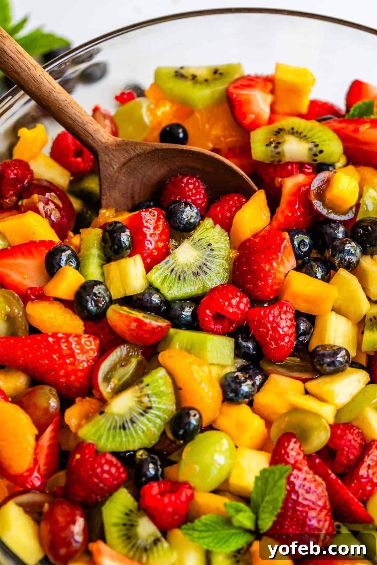 A wooden spoon delicately scooping a portion of glistening fruit salad, coated with a bright glaze, from a clear glass bowl, showcasing the refreshing mix of fruits.