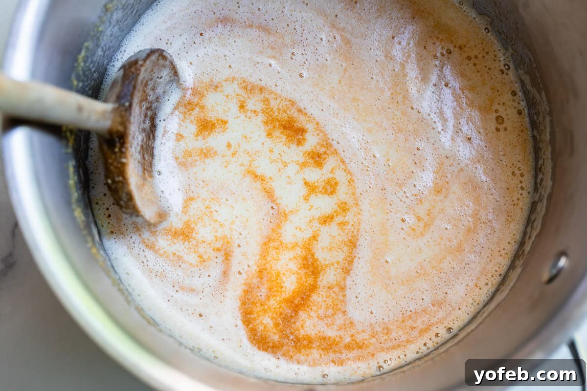 The Ultimate No Bake Cheesecake 22 wooden spoon stirring through browned butter with foam on top in metal pan.