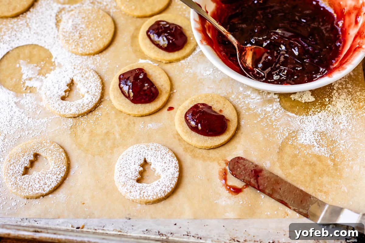 Melt-in-Your-Mouth Linzer Delights 17 A tray filled with unassembled Linzer cookies, featuring plain bottoms, powdered sugar-dusted tops with cutouts, and a bowl of jam ready for filling.