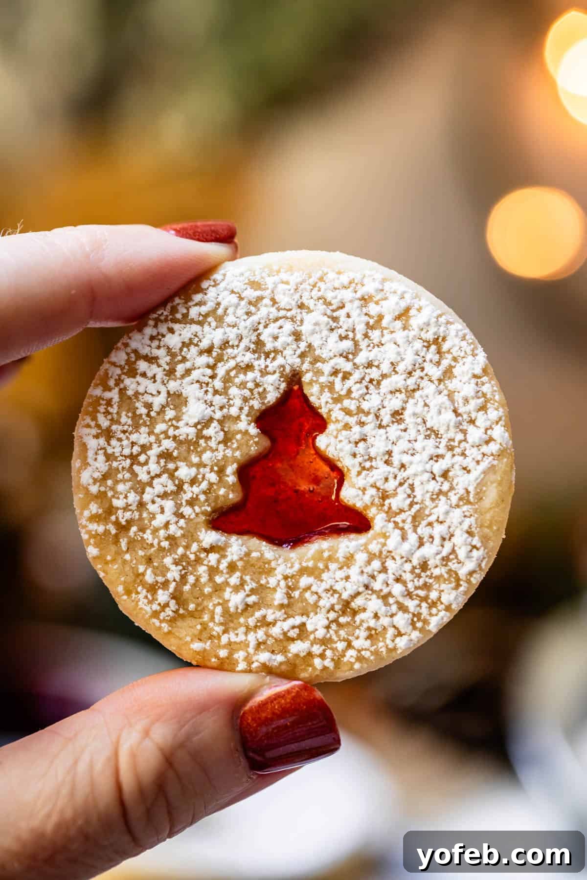 Melt-in-Your-Mouth Linzer Delights 5 A hand gently holding a Linzer cookie, featuring a whimsical Christmas tree cutout that reveals a rich, red jam filling.