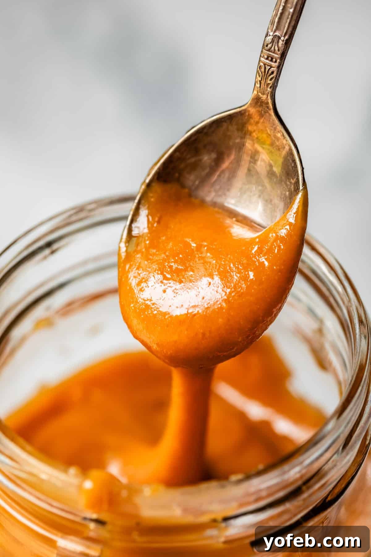 Dreamy Salted Caramel Sauce 5 A metal spoon drips thick, creamy salted caramel sauce into a glass jar, showcasing its rich, luxurious consistency.