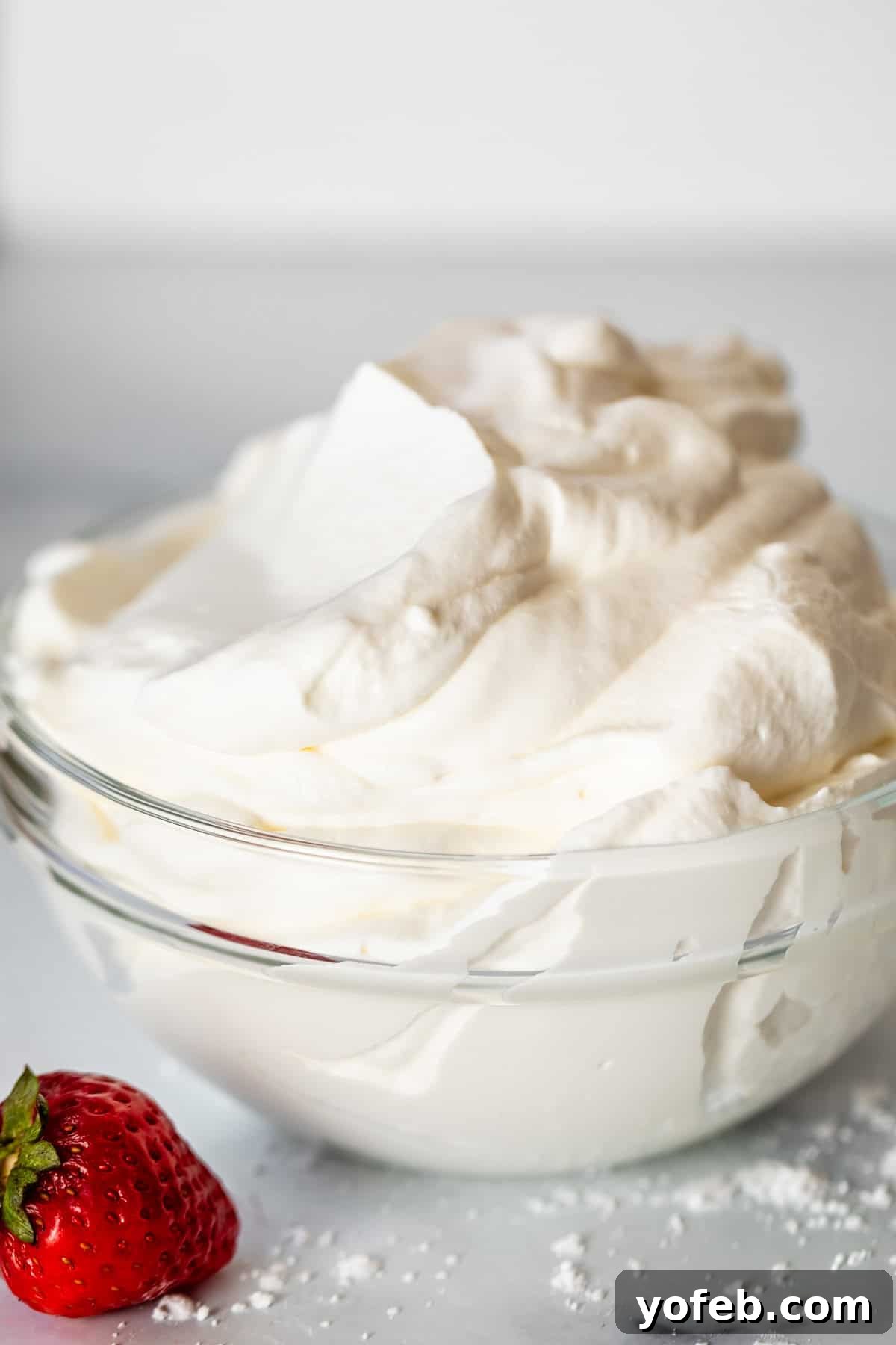 Homemade Whipped Cream Made Easy 21 glass bowl of fluffy whipped cream on a counter with a single strawberry next to it.