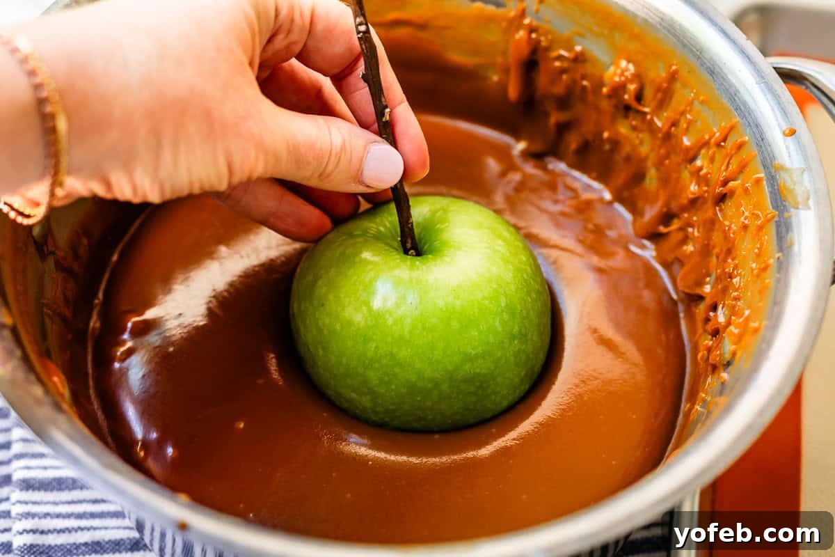 My Ultimate Caramel Apple 22 A hand gently lowering a green apple, held by its stick, into a large pot of luscious, hot caramel, illustrating the dipping process.