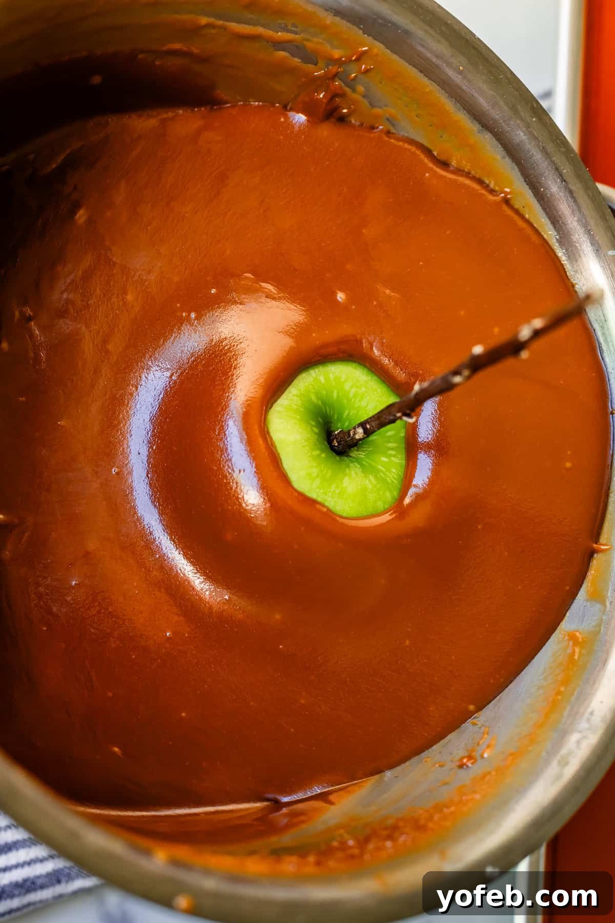 My Ultimate Caramel Apple 24 A metal pan tilted, with an apple on a stick completely submerged in the thick, glossy caramel, ensuring full coverage.