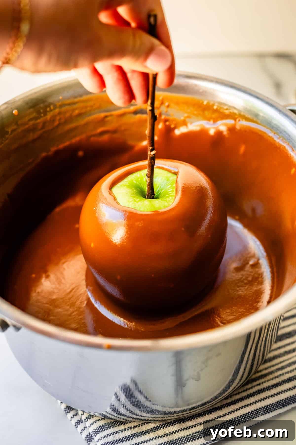 My Ultimate Caramel Apple 25 A hand carefully lifts a fully caramel-coated Granny Smith apple from the pot, showcasing its smooth, glistening exterior.