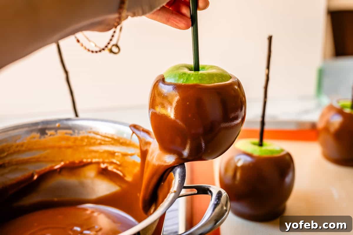 My Ultimate Caramel Apple 27 A hand gently scrapes excess caramel from the bottom edge of a dipped apple against the side of the pan, ensuring a clean finish.