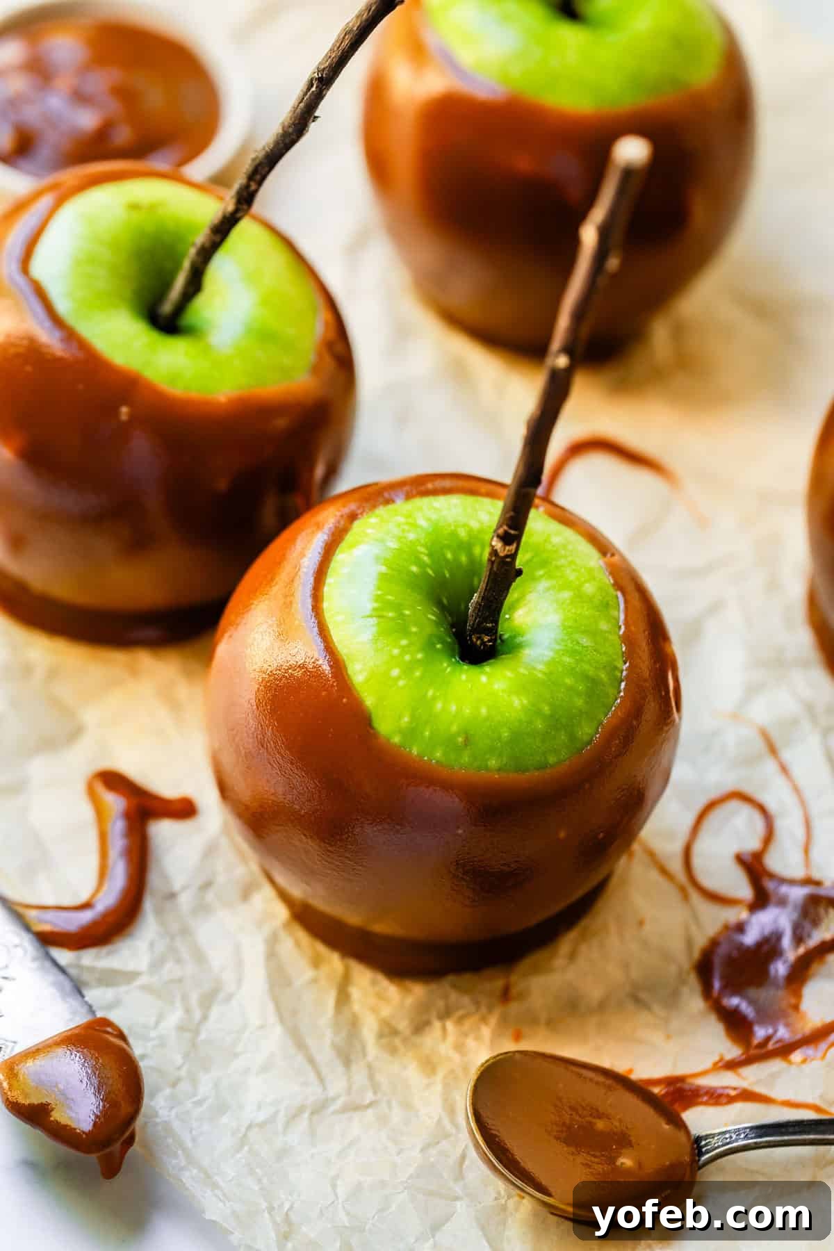 My Ultimate Caramel Apple 35 A pristine green caramel apple with a natural twig stick, accompanied by a spoon drizzled with luscious caramel, all resting on parchment paper.