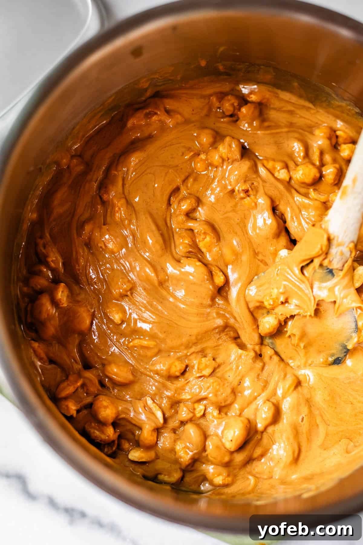 Golden Peanut Snap 13 A saucepan filled with a thick, creamy, caramel-colored mixture and an abundance of peanuts is being stirred with a white spatula on a pristine marble countertop, ready for the next stage of preparation.