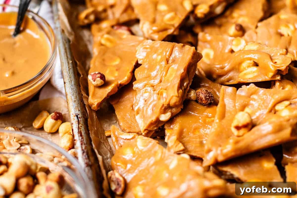 Golden Peanut Snap 17 Close-up of golden, crunchy peanut brittle pieces stacked on a tray, revealing visible whole peanuts embedded in the caramelized candy. A glass bowl of creamy peanut butter and extra peanuts are also visible nearby, emphasizing the nutty theme.