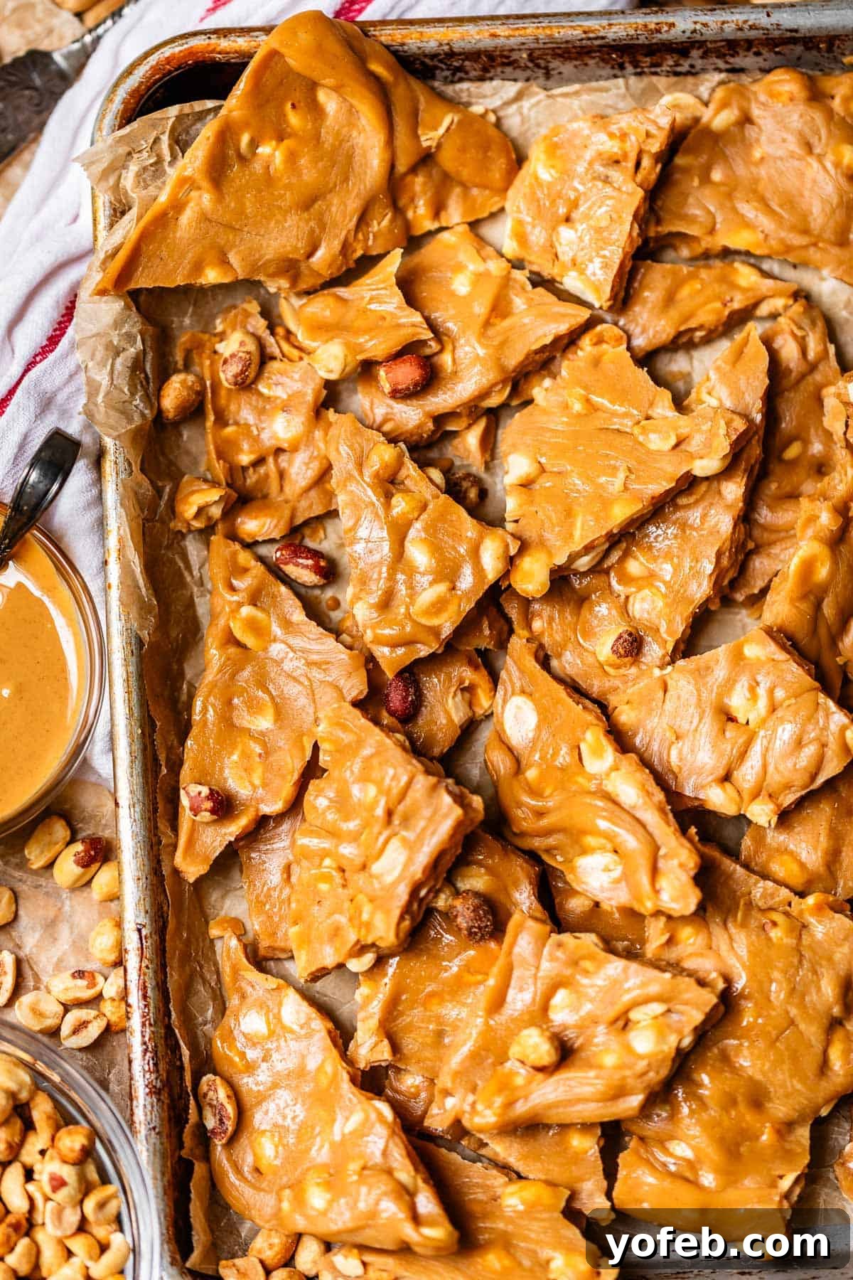 Golden Peanut Snap 3 A baking tray lined with parchment paper showcases broken pieces of golden brown peanut brittle, brimming with visible peanuts. A small bowl of creamy peanut butter and scattered whole peanuts rest beside the tray.