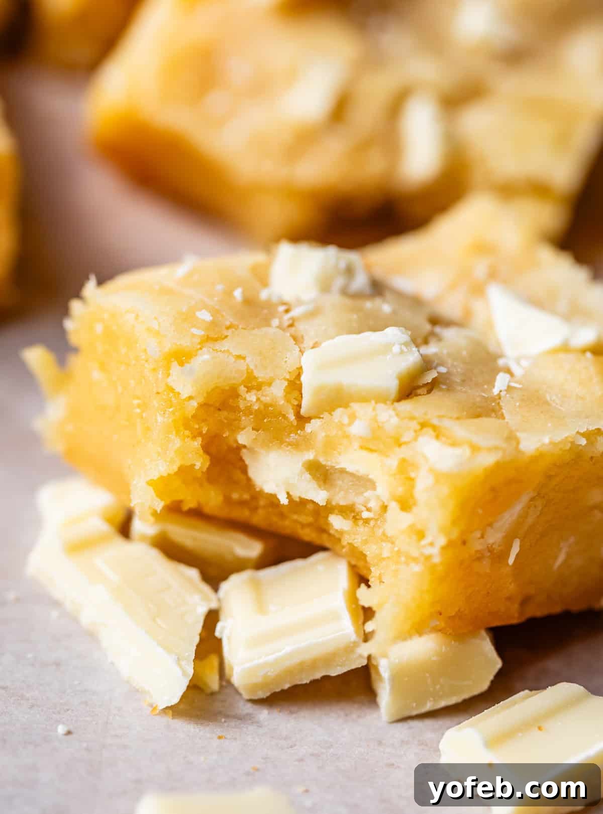 Irresistible White Chocolate Brownies 2 A piece of white chocolate brownie with a bite taken, sitting on chopped white chocolate. The brownie is rich and fudgy, with a perfect golden-brown crust.