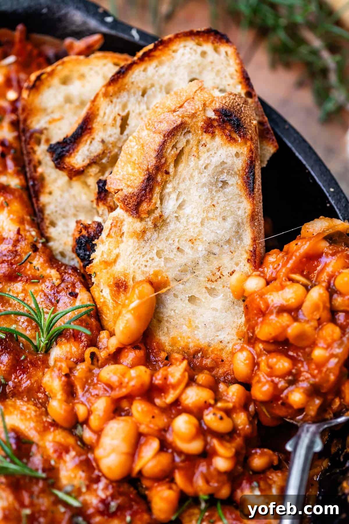 Rustic Tomato, White Bean & Cheese Bake 15 A cast iron skillet of cheesy white bean tomato bake, garnished with fresh herbs and three inviting slices of sourdough bread, one with a metal spoon, ready to be enjoyed.