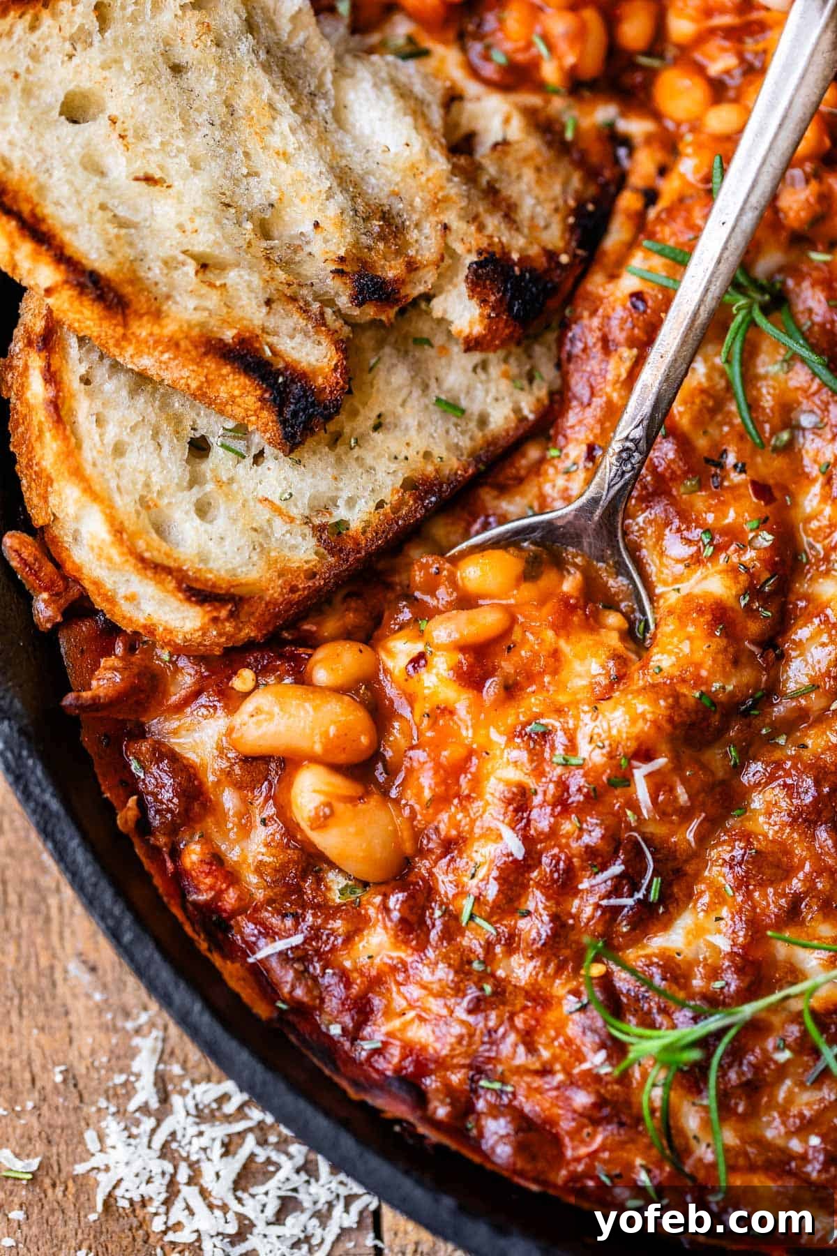Rustic Tomato, White Bean & Cheese Bake 16 A cozy scene of a metal spoon and a slice of sourdough bread nestled into a cast iron skillet of the cheesy white bean tomato bake, ready to be savored.