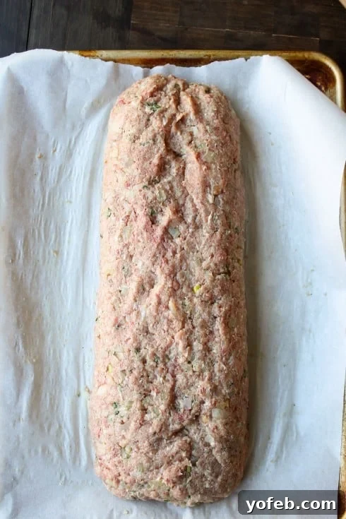 Ina Garten's 1770 House Meatloaf 8 A perfectly shaped raw meatloaf, molded into a smooth cylinder, resting on a parchment-lined baking sheet, prepared for baking.