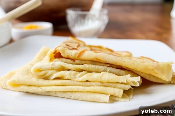 Zesty Meyer Lemon & Cardamom Crepes 3 folded crepes stacked on white plate with powdered sugar and lemon zest in the background.