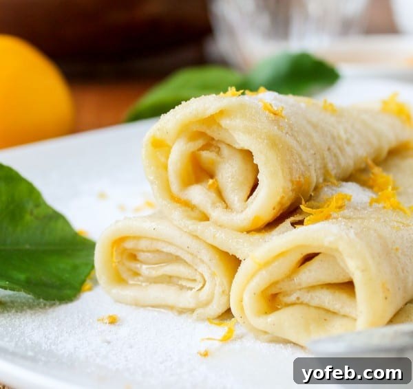 Zesty Meyer Lemon & Cardamom Crepes 4 stack of three rolled crepes on white plate topped with sifted powdered sugar and lemon zest.