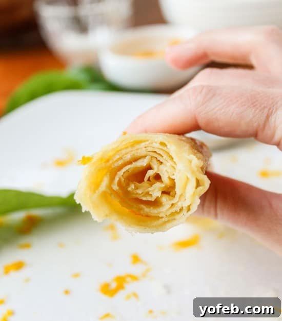 Zesty Meyer Lemon & Cardamom Crepes 5 hand holding one rolled-up crepe with bite taken out of it over plate with powdered sugar and lemon zest.