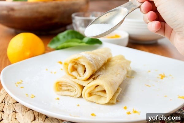 Zesty Meyer Lemon & Cardamom Crepes 6 pouring sugar on rolled up crepes stacked on white plate on wooden table with lemon and toppings in the background.
