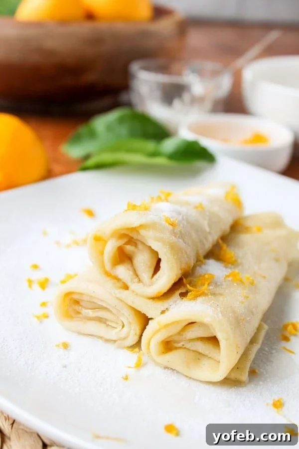 Zesty Meyer Lemon & Cardamom Crepes 7 three crepes stacked and rolled up on white plate in vertical photo with meyer lemons, leaves, and bowls with toppings in background.