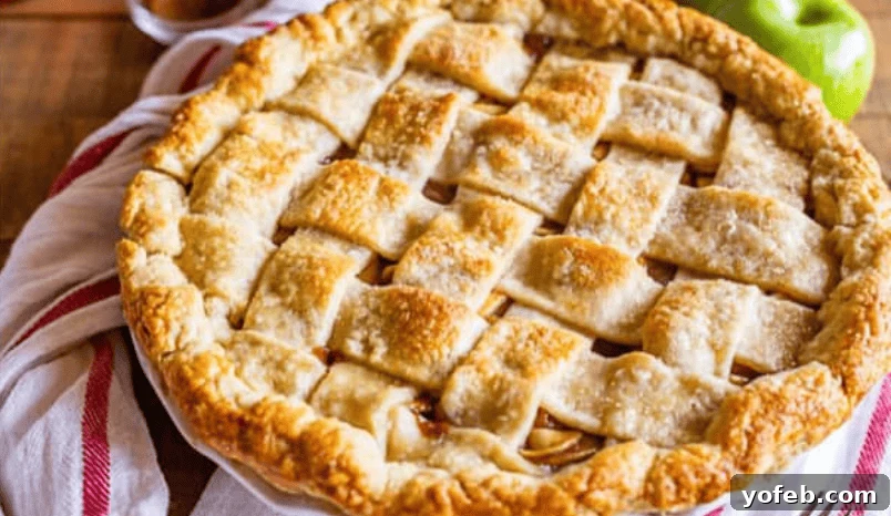 Crafting Your Dream Apple Pie and Apple Crisp 2 A freshly baked apple pie with a golden brown, lattice crust sits on a white dish, with a red-and-white striped towel and a green apple in the background.
