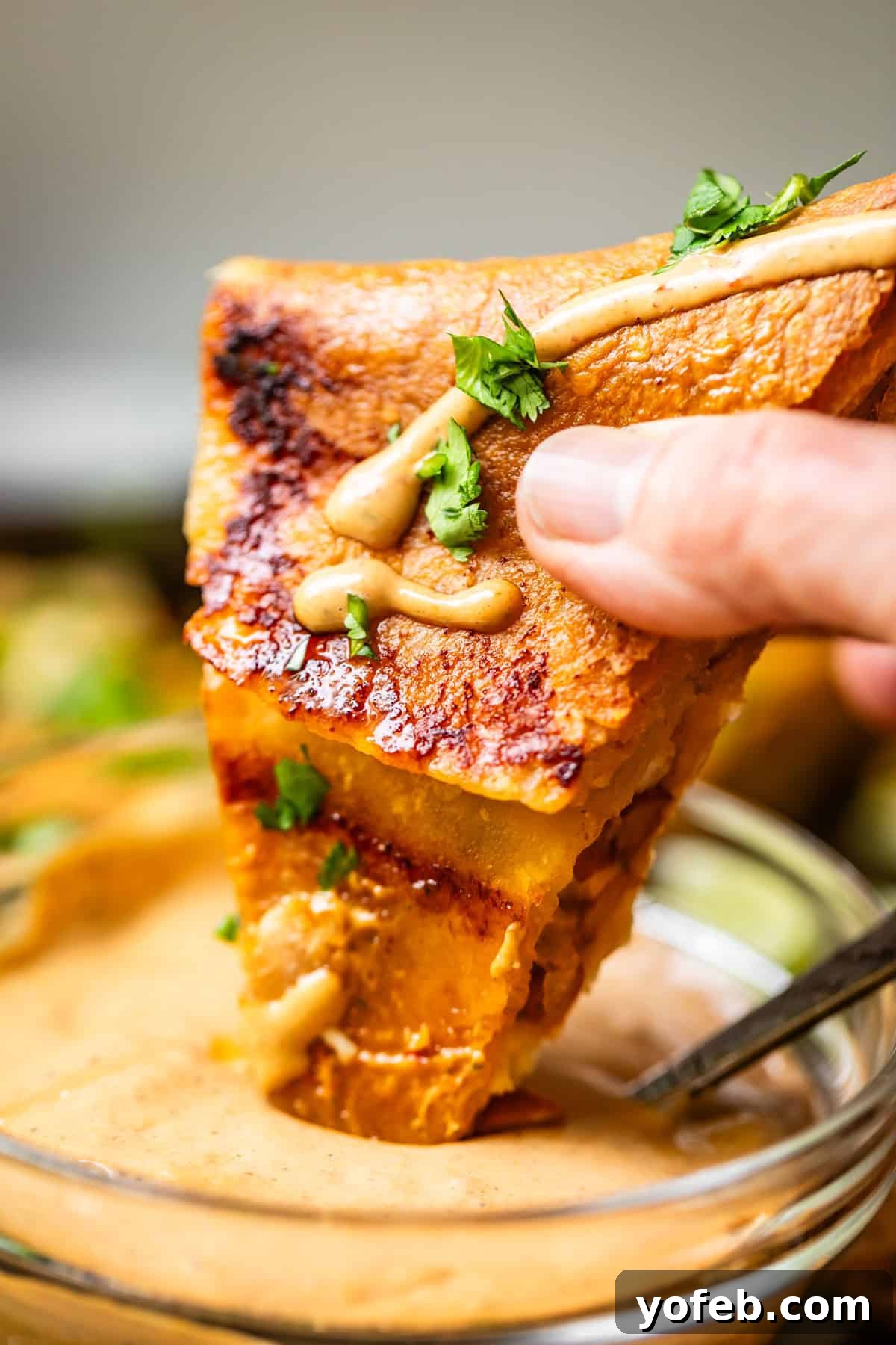 Crispy Sheet Pan Chicken Quesadillas 5 A hand holds a slice of cheesy quesadilla topped with herbs and a sauce drizzle, dipping it into a bowl of creamy dipping sauce.